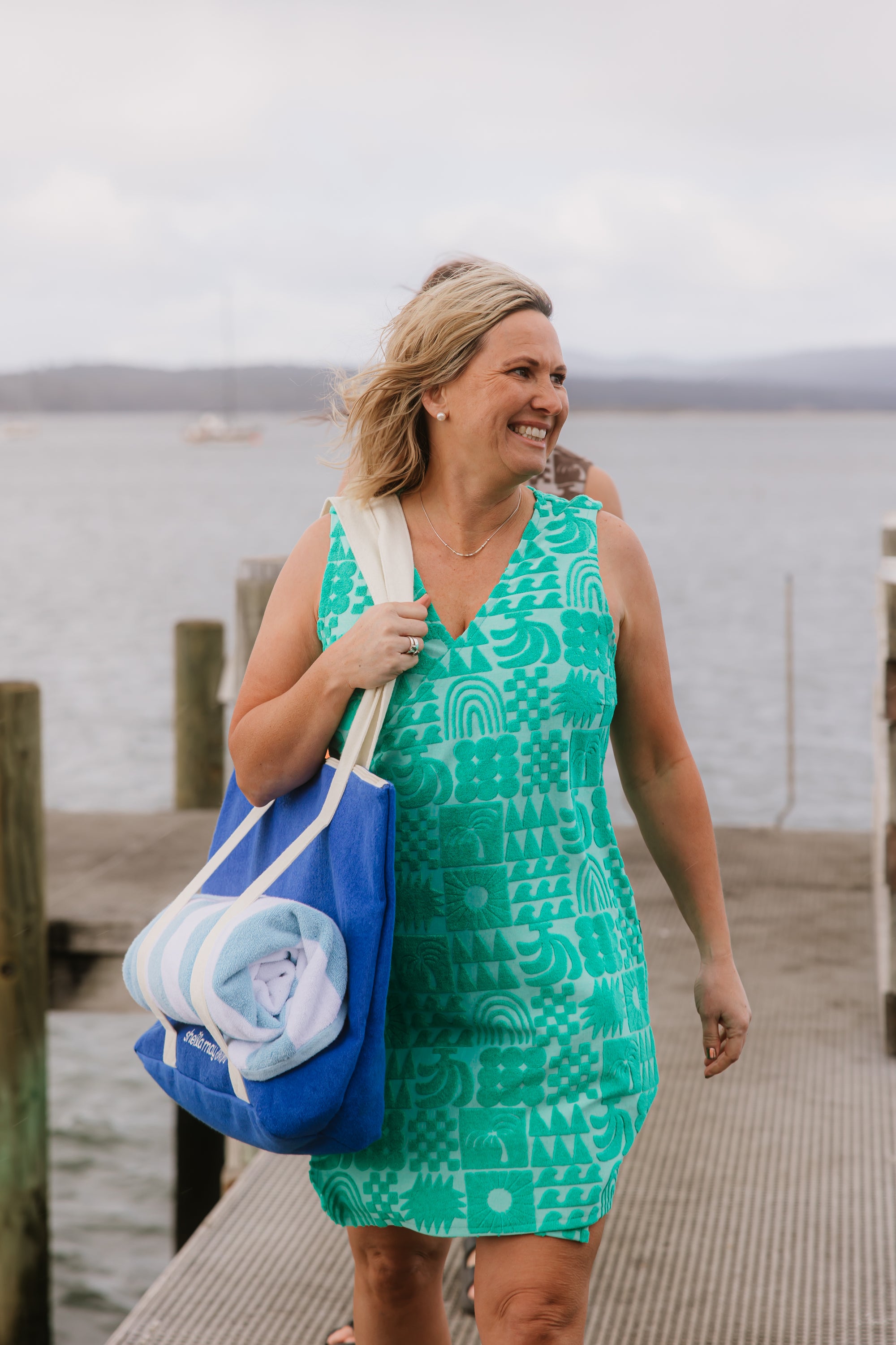 Woman in a sprout green terry wrap beach dress holding a blue tote bag on a dock by water.